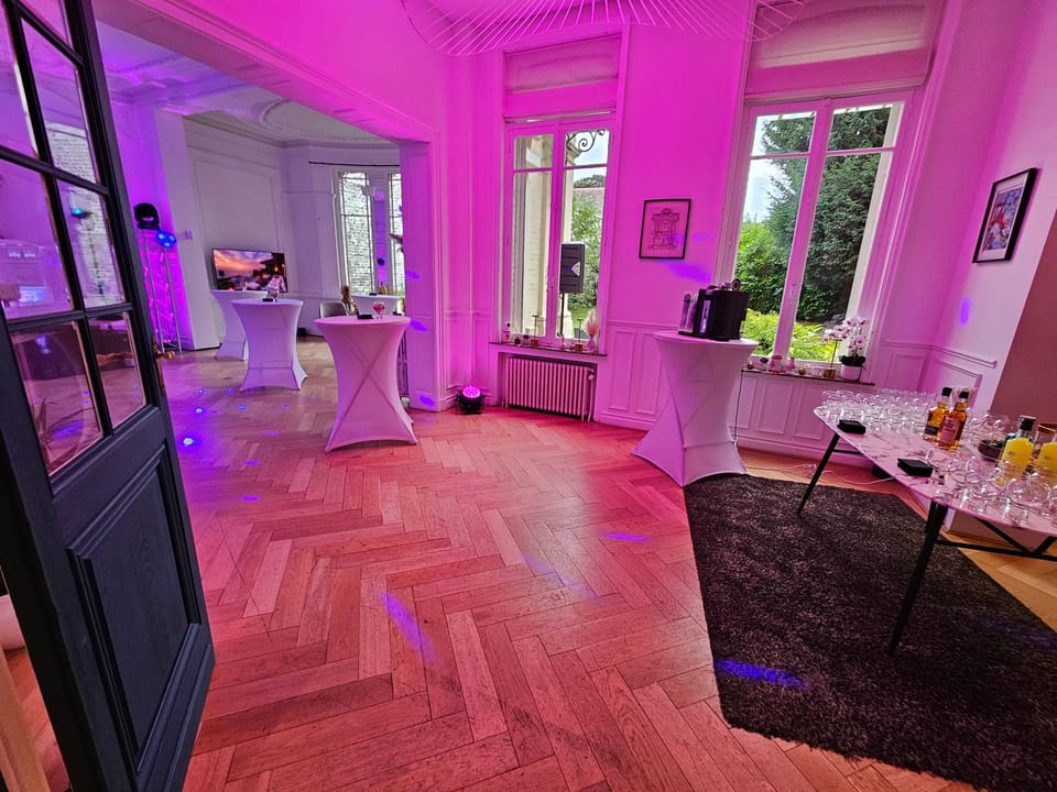 Salon aux lumières roses et violettes avec mange-debout, parquet en chevrons et table de boissons, prêt pour la soirée