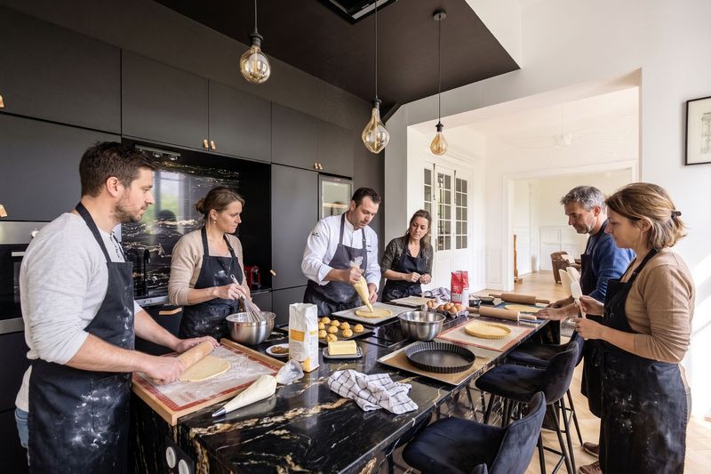 Équipe en tablier autour de l'îlot en marbre de la cuisine du 119 : un chef montre comment garnir une tarte, pâte, œufs et poches à douille sur le plan de travail